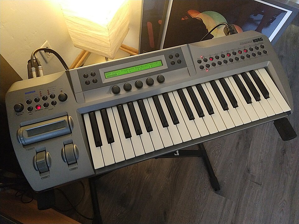 A silver Korg Prophecy 37-key monophonic synthesizer on a stand, with its green display lit and a ribbon controller and two wheels on the left.
